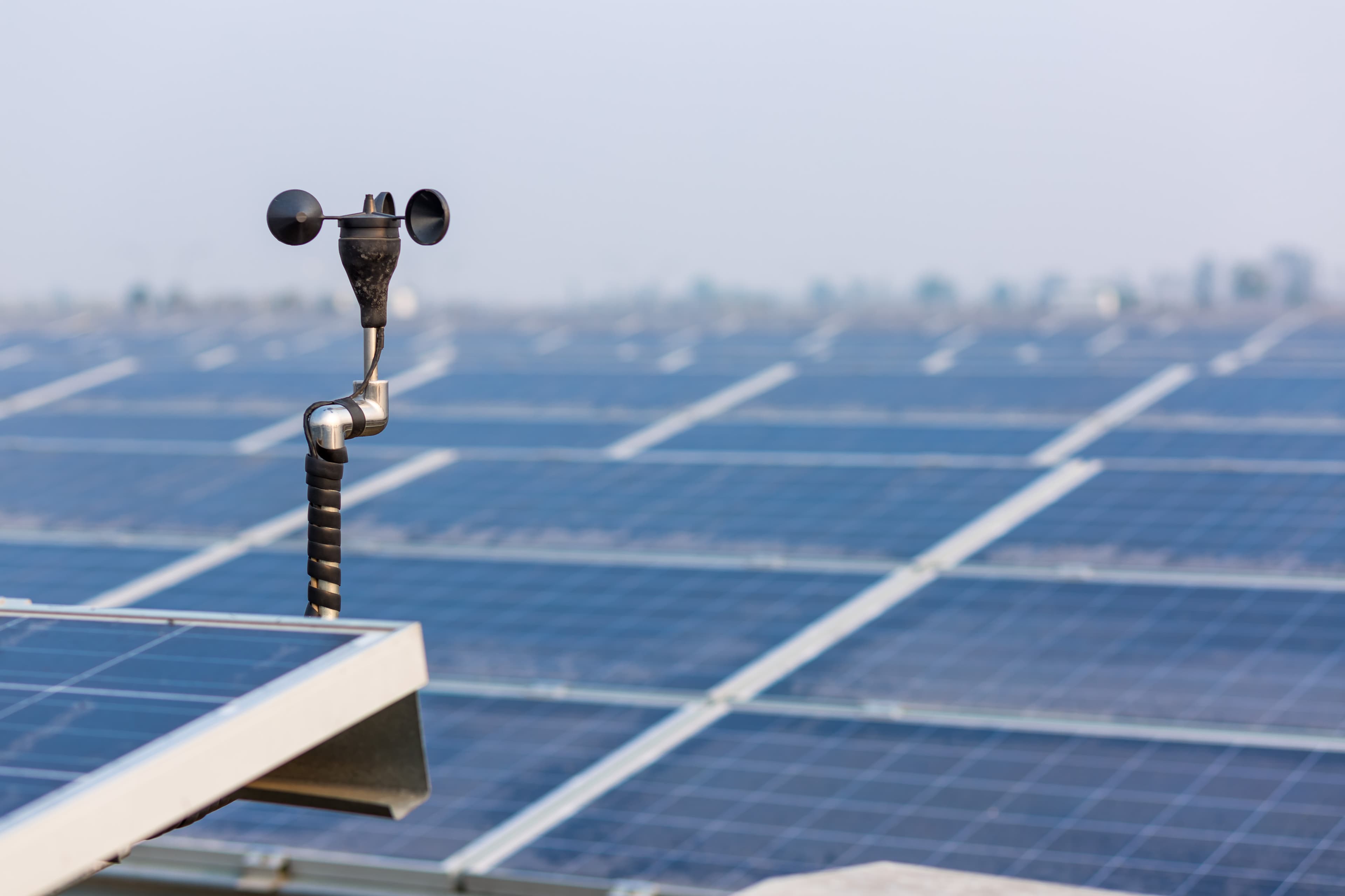 Nahaufnahme eines Windmessers auf einem Dach, im Hintergrund sind Solarmodule und ein dunstiger Himmel zu sehen.