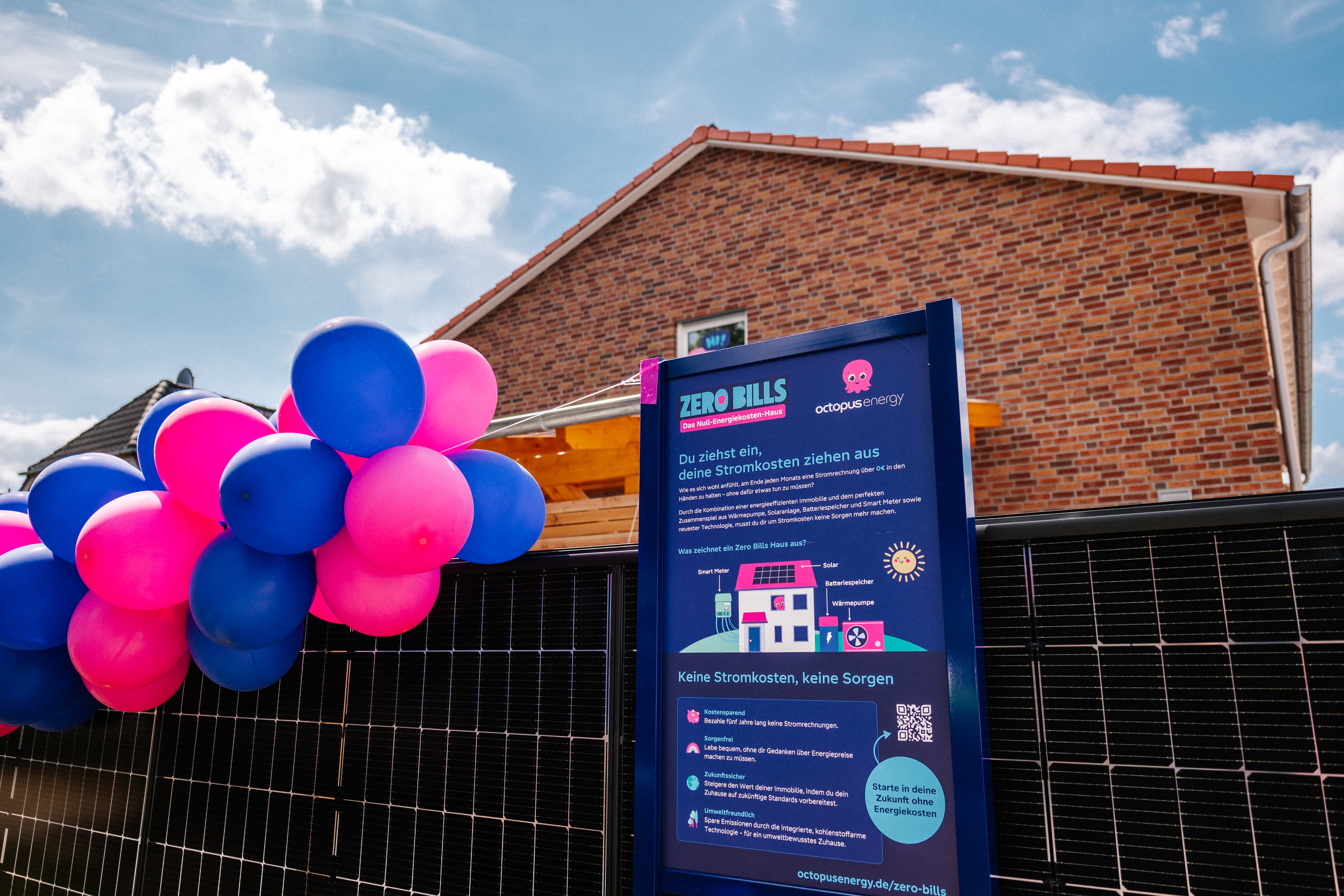 Schild mit der Aufschrift „Zero Bills“ vor einem Haus mit Sonnenkollektoren, bunten Luftballons und einem Backsteinhaus im Hintergrund unter blauem Himmel.