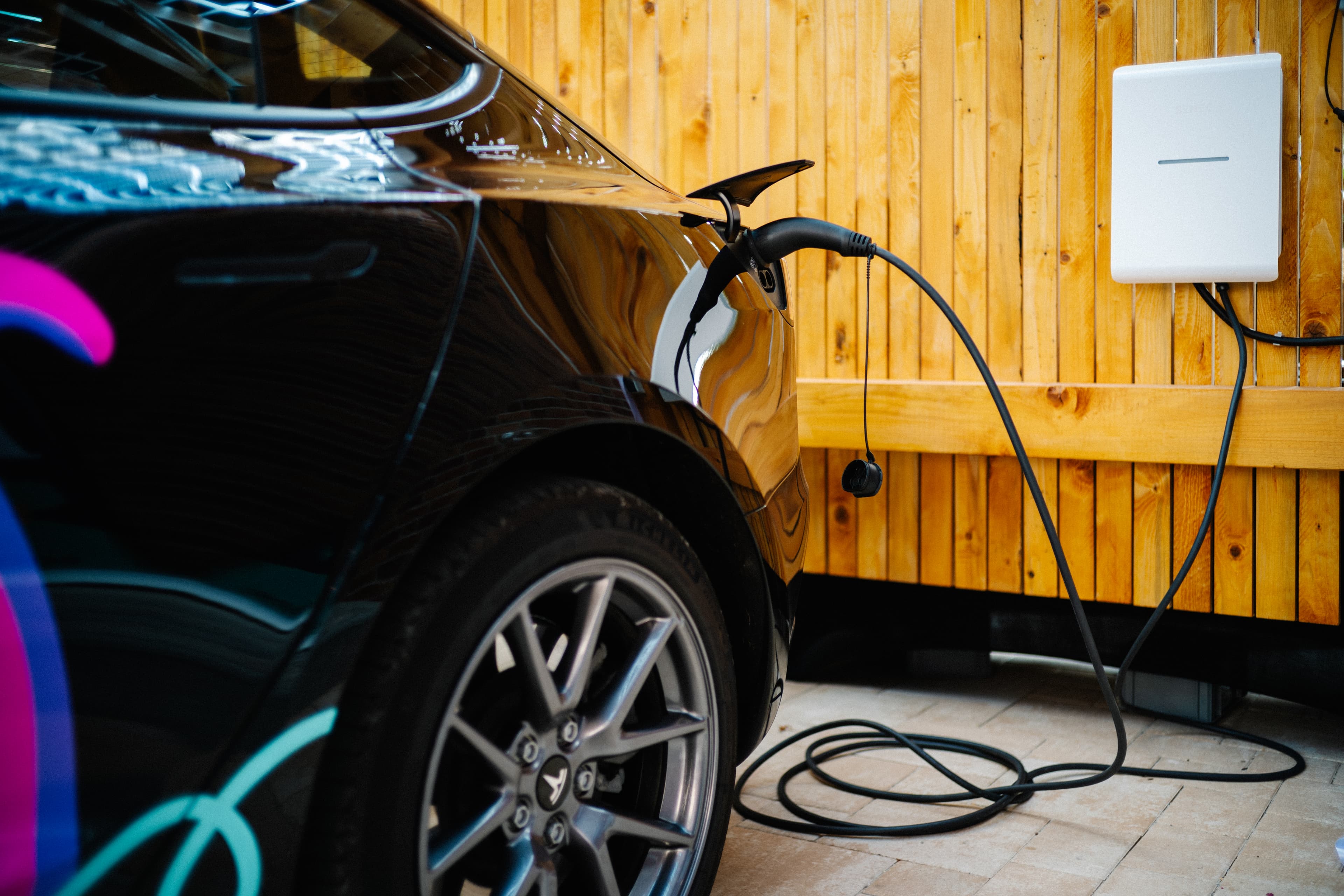 Ein schwarzes Elektroauto wird gerade aufgeladen; es ist an eine an der Wand montierte Ladestation angeschlossen, im Hintergrund ist eine Holzvertäfelung zu sehen.