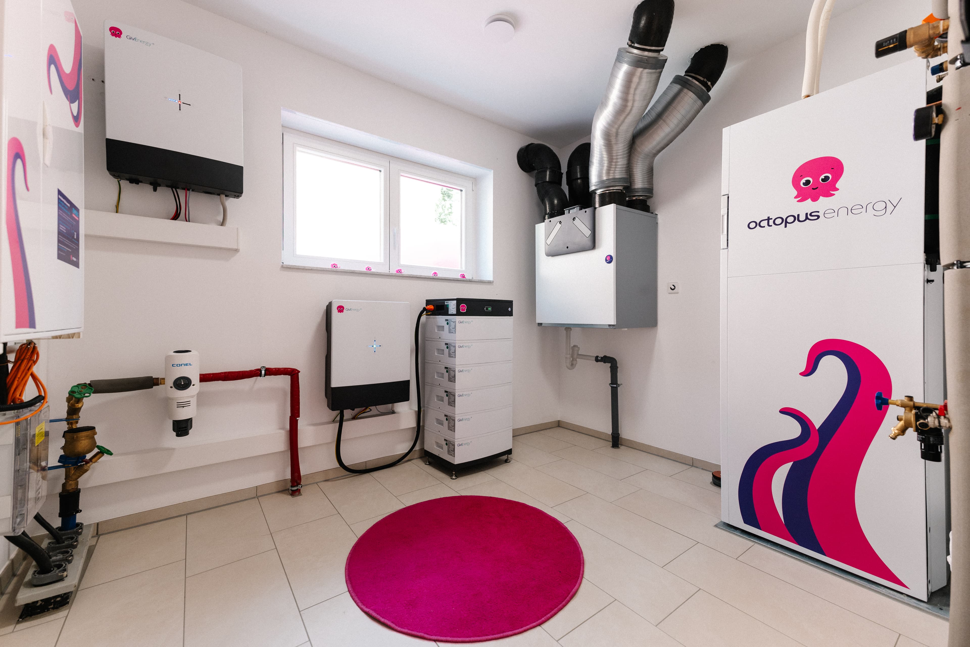 Moderner Hauswirtschaftsraum mit Geräten von Octopus Energy, Rohrleitungen, einem Batteriesystem und einem leuchtend pinkfarbenen, runden Teppich auf einem Fliesenboden.