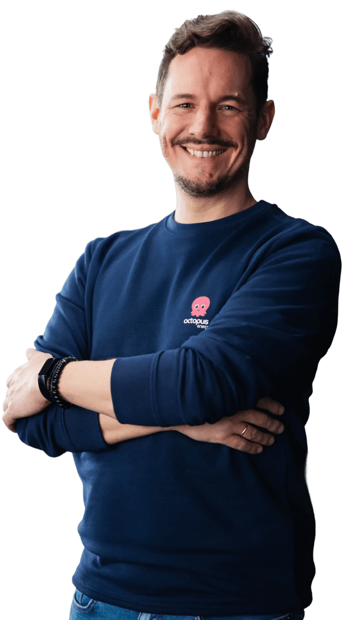 Smiling man in a blue sweatshirt with an octopus logo, arms crossed, wearing a watch and wedding band.